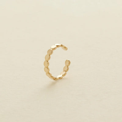 A delicate gold cuff earring made up of round flat discs laying side by side photographed on a clean, light beige background.