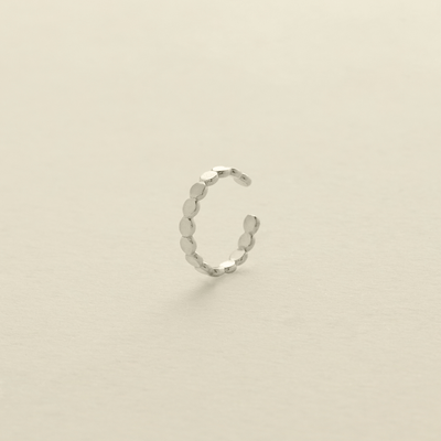 A delicate silver cuff earring made up of round flat discs laying side by side photographed on a clean, light beige background.