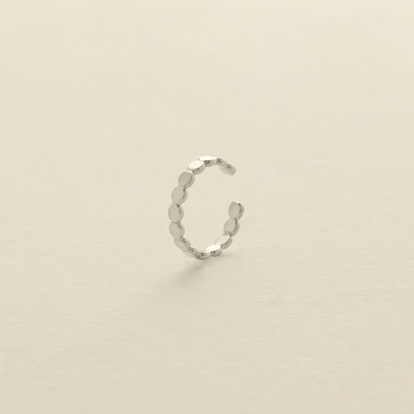 A delicate silver cuff earring made up of round flat discs laying side by side photographed on a clean, light beige background.
