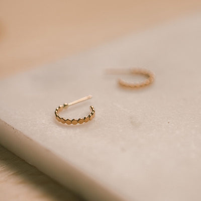 A textured gold hoop with a straight post, laying on a white marble surface with a blurred background.