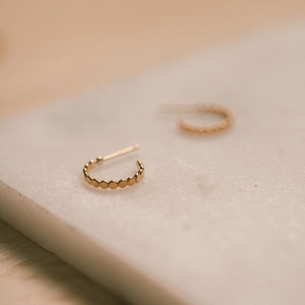 A textured gold hoop with a straight post, laying on a white marble surface with a blurred background.
