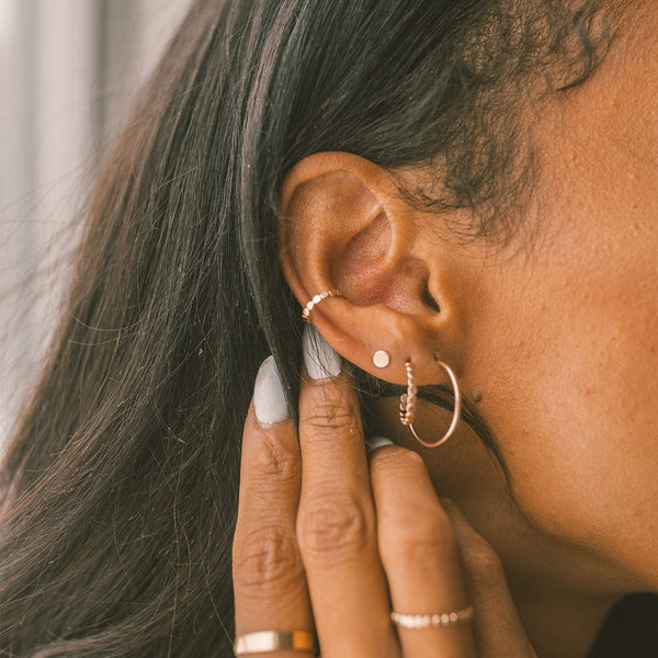 A close-up of a person's ear wearing a large thin gold hoop, a thicker gold hoop made of small round flat discs, a gold flat disc stud, and a textured gold ear cuff.