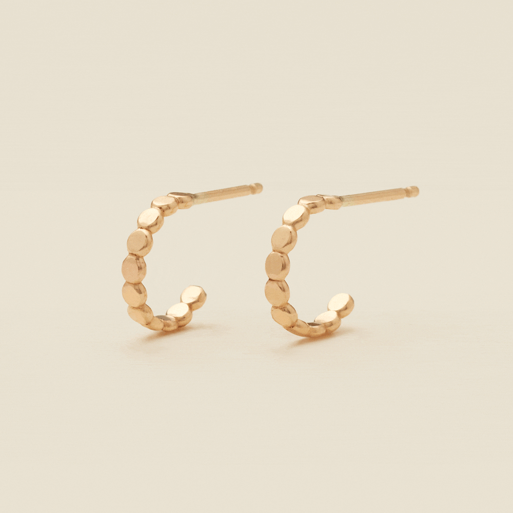 A gif of two gold hoop earrings with a straight post, made up of round flat discs side by side, set against a solid beige background.