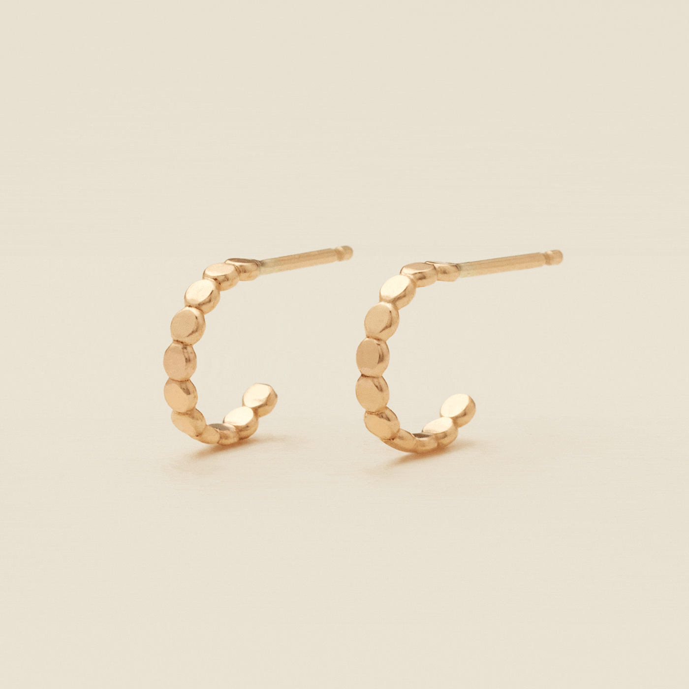 A gif of two gold hoop earrings with a straight post, made up of round flat discs side by side, set against a solid beige background.
