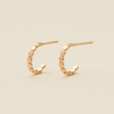 A gif of two gold hoop earrings with a straight post, made up of round flat discs side by side, set against a solid beige background.