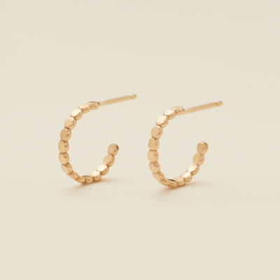 A gif of two gold hoop earrings with a straight post, made up of round flat discs side by side, set against a solid beige background.