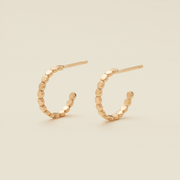 A gif of two gold hoop earrings with a straight post, made up of round flat discs side by side, set against a solid beige background.
