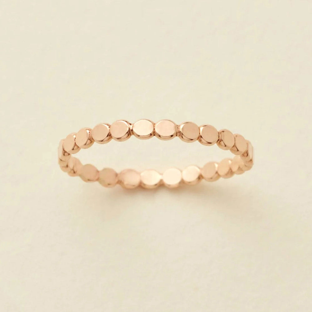 A delicate gold eternity band ring featuring small, flat circular discs, photographed on a clean, solid, warm off-white background.