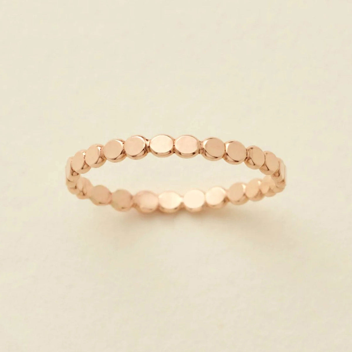 A delicate gold eternity band ring featuring small, flat circular discs, photographed on a clean, solid, warm off-white background.