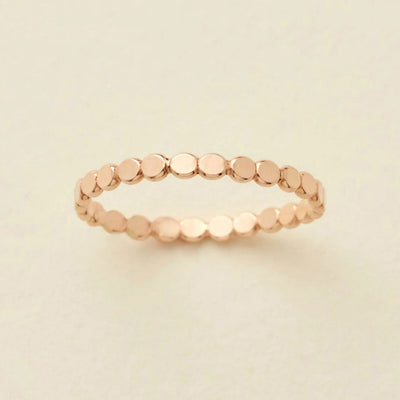 A delicate gold eternity band ring featuring small, flat circular discs, photographed on a clean, solid, warm off-white background.