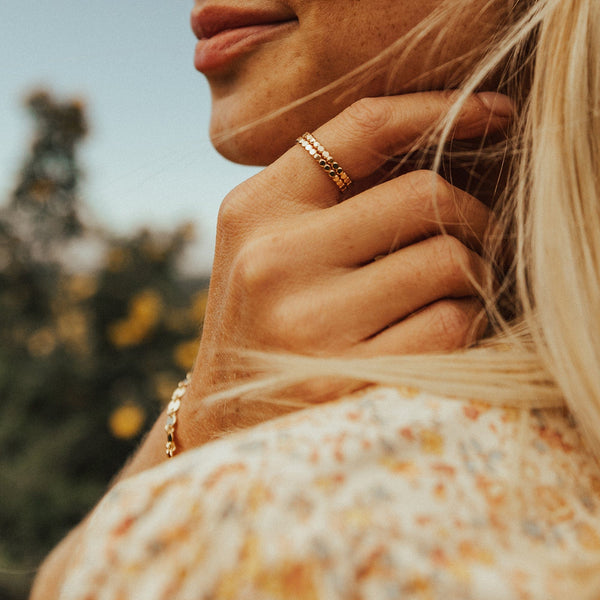 A close-up image of a person's hand resting near their face, showcasing two stacked gold rings comprised of round flat discs set side by side, set with a blurred outdoor background.