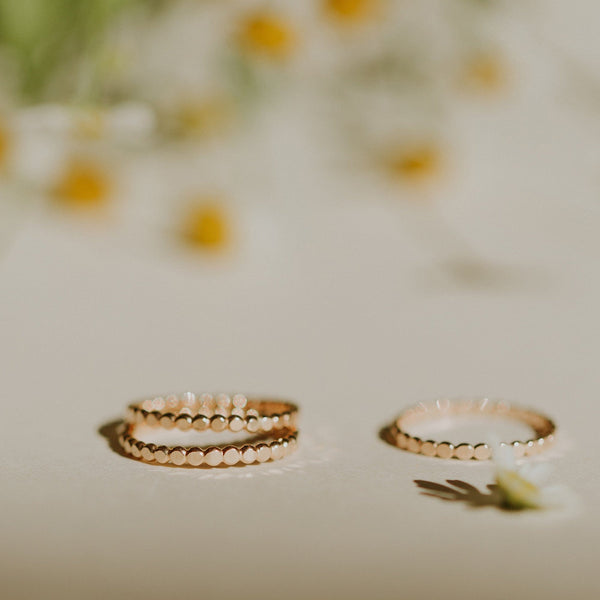 A set of three gold eternity rings with a band of small flat round discs, set on a light beige background with blurred yellow flowers in the background.