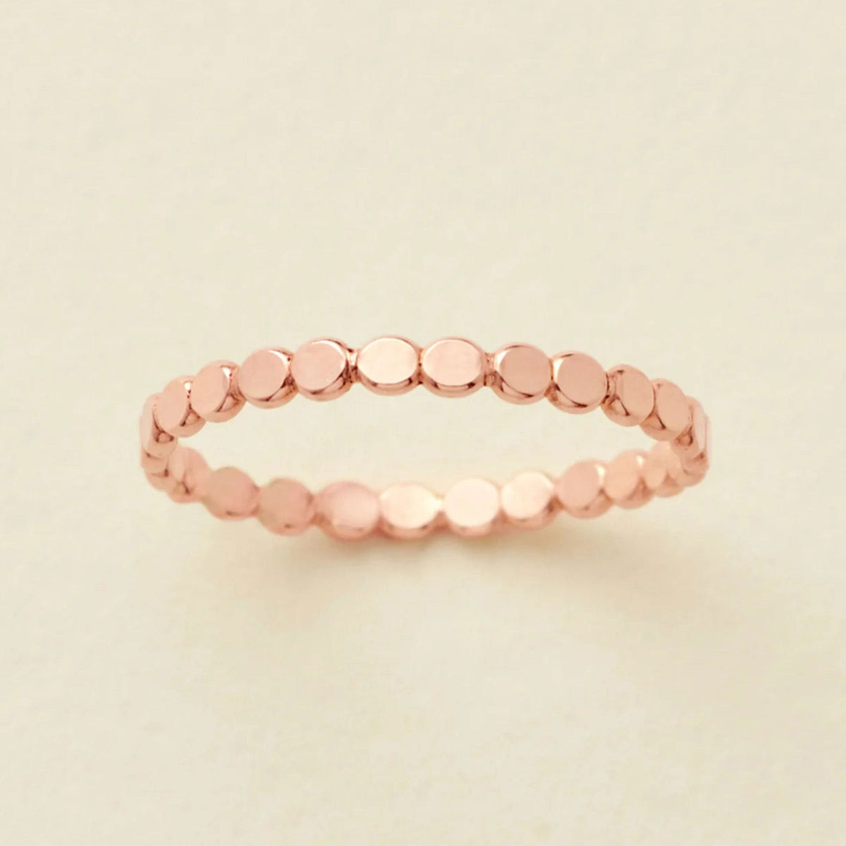 A thin rose gold band ring made of small, flat, circular disc-like links, photographed on a light beige background.