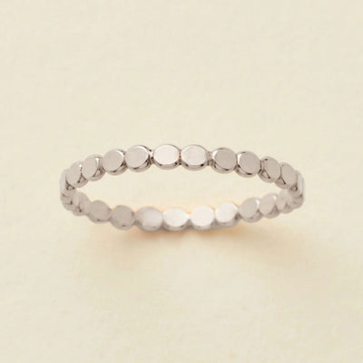A delicate silver eternity band ring featuring small, flat circular discs, photographed on a clean, solid, warm off-white background.