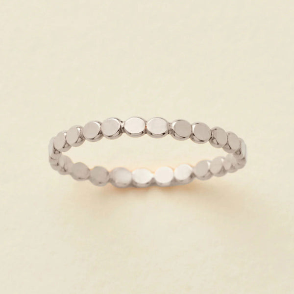 A delicate silver eternity band ring featuring small, flat circular discs, photographed on a clean, solid, warm off-white background.