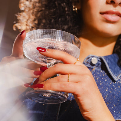 A close-up of a woman holding a crystal martini glass, showcasing a gold eternity style ring and single pearl gold ring, paired with a jean jacket.