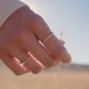 A short video of a person's hand walking through a field and touching the beige plants, showcasing a gold thin band ring with a single pearl and a gold eternity style ring with round bezel-set clear gemstones.