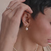 A close-up video of a person's ear wearing a gold round stud earring with a pearl hanging from the bottom, and a clear gemstone prong set stud, set against a beige background.