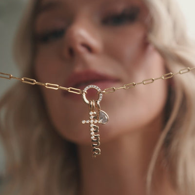 A short video of a woman holding and wearing a gold paperclip link chain necklace with a round ring in the center covered in clear accent stones. On the ring are a gold cross charm with clear gemstones, a vertical charm spelling out 'mama' in script font, and a clear pear shaped gemstone charm.