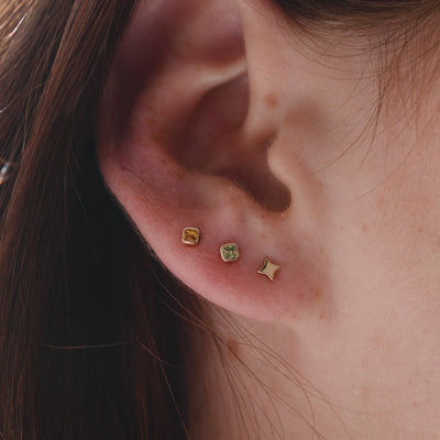 A short close-up video of a person's ear wearing a gold four point star stud and two square bezel-set gemstone studs, one with an amber stone and one with a light green stone.