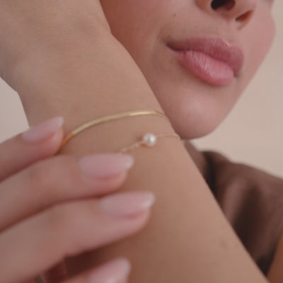 A close-up of a persons wrist wearing a flat gold bracelet and a dainty gold chain bracelet with a single pearl in the center, set against a beige background.