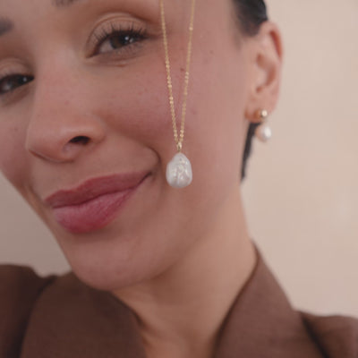 A close-up video of a woman holding a dainty gold pendant chain with a large organic pearl charm, then putting the necklace on.