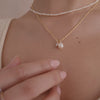 A close-up video of a person's neck wearing a pearl beaded strand choker necklace layered with a longer textured gold pendant chain with a pearl charm and a square clear gemstone charm.