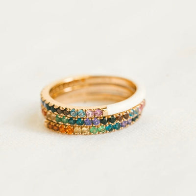 A stack of three gold rings, with the back half of the ring covered in white enamel and the front half being a single row of colored gemstones in a rainbow sequence, shown on a solid white surface.