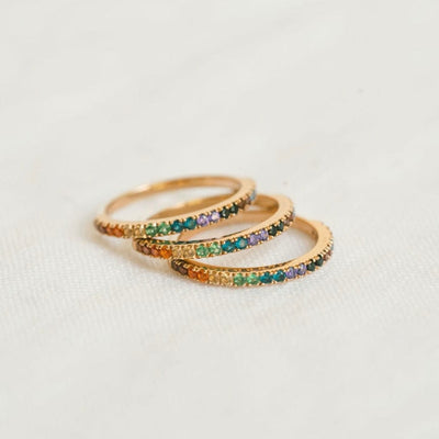 A stack of three gold rings, each with a single row of colored gemstones in a rainbow sequence across the front, shown on a textured white surface.