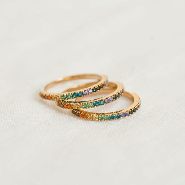 A stack of three gold rings, each with a single row of colored gemstones in a rainbow sequence across the front, shown on a textured white surface.