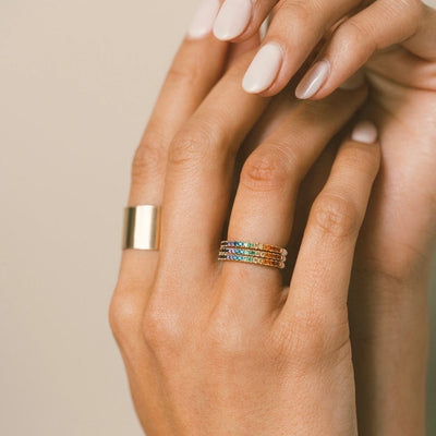 A person's hands wearing a stack of three gold rings, each with a single row of a rainbow of colored gemstones, shown on a light beige background.