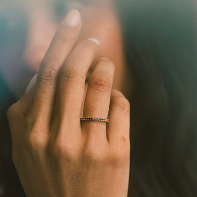 A close-up of a person's finger wearing a gold ring with a single row of colored gemstones in a rainbow sequence, symbolizing pride.