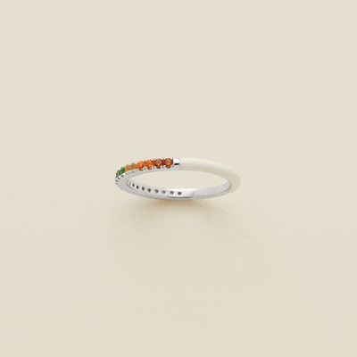 A gif of a silver ring, showing different angles of the ring in a 360 degree motion. The back half of the ring is covered in white enamel, and the front has a single row of small colored gemstones in a rainbow pattern. The ring is shown on a solid light beige background.