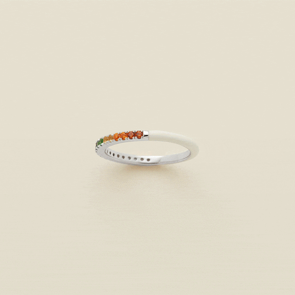 A gif of a silver ring, showing different angles of the ring in a 360 degree motion. The back half of the ring is covered in white enamel, and the front has a single row of small colored gemstones in a rainbow pattern. The ring is shown on a solid light beige background.