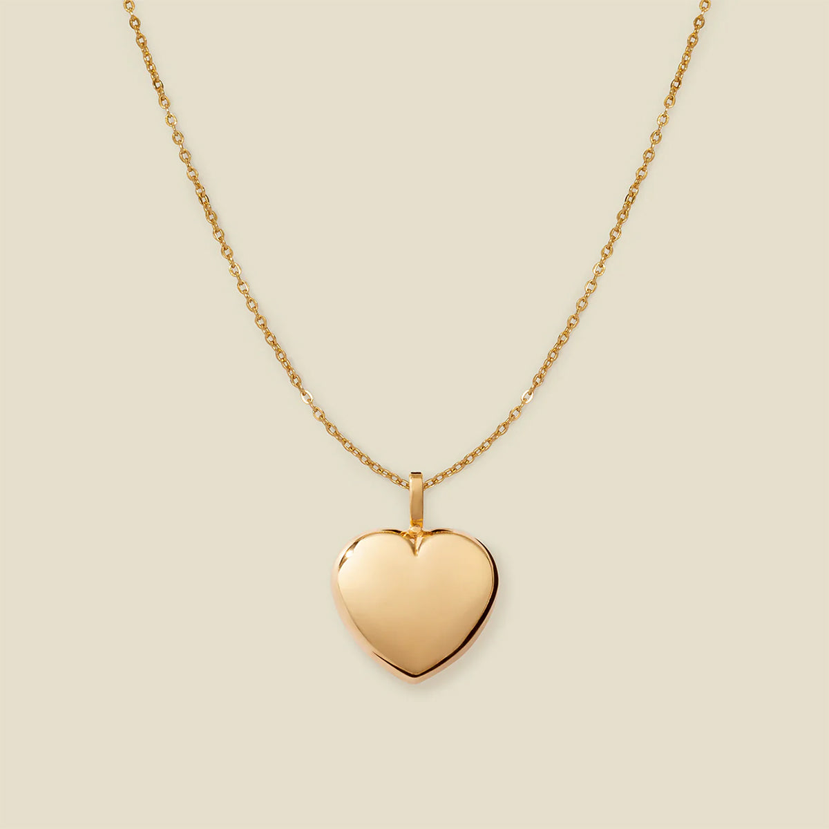A polished gold heart-shaped pendant necklace on a delicate gold chain, centered against a solid light beige background.