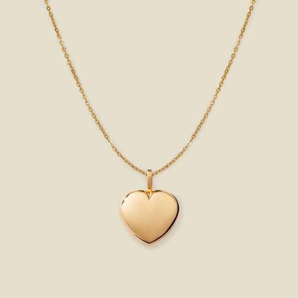 A polished gold heart-shaped pendant necklace on a delicate gold chain, centered against a solid light beige background.