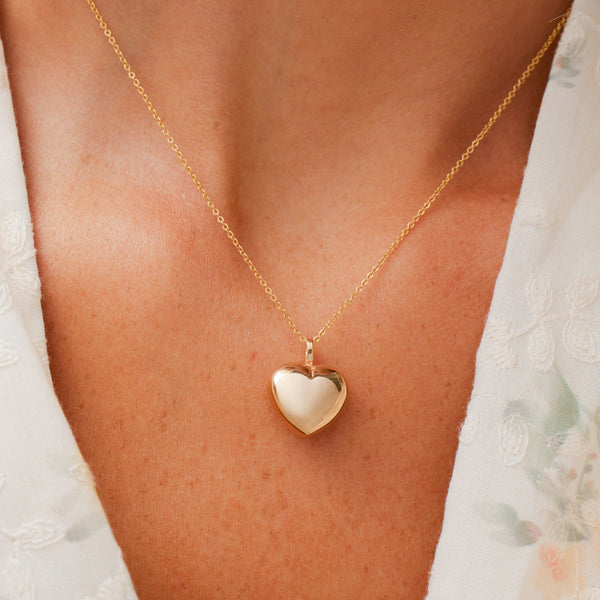 A close up of a person's chest wearing a delicate gold chain necklace with a large polished heart pendant.