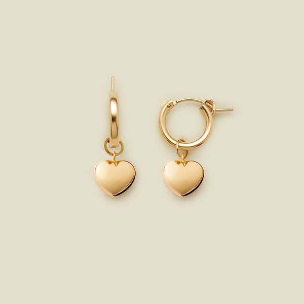 A pair of gold huggie hoop earrings with dangling polished heart charms, displayed on a solid light beige background.