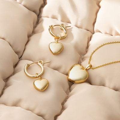 A pair of gold huggie hoop earrings with dangling polished heart charms and a delicate chain necklace with a larger, matching heart charm, shown on a light beige puffy quilted blanket.