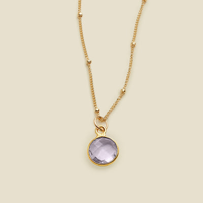 A gold chain pendant necklace with stationary gold beads, with a light purple gemstone charm, set against a solid beige background.