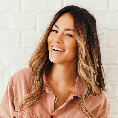 A woman in a light maroon corduroy button up top wearing a delicate gold pendant necklace with two vertical bar charms in varying lengths.