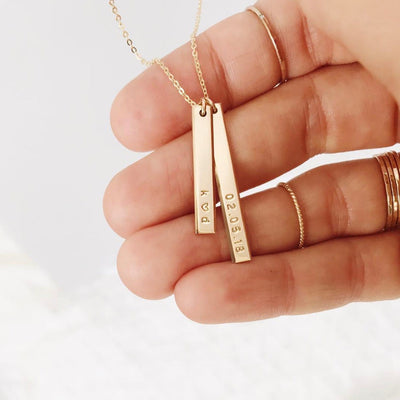 A person's hand wearing multiple gold rings holding the the charms of a gold pendant chain necklace. There are two vertical bar charms in different lengths, and both are engraved with either 'k ♥ d' or '02.05.18'.