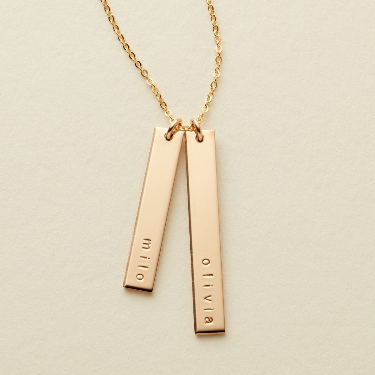 A delicate gold name necklace featuring two vertical bar pendants engraved with the names 'milo' and 'olivia', suspended from a delicate chain on a light beige background.