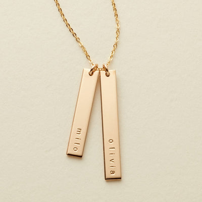 A delicate gold name necklace featuring two vertical bar pendants engraved with the names 'milo' and 'olivia', suspended from a delicate chain on a light beige background.