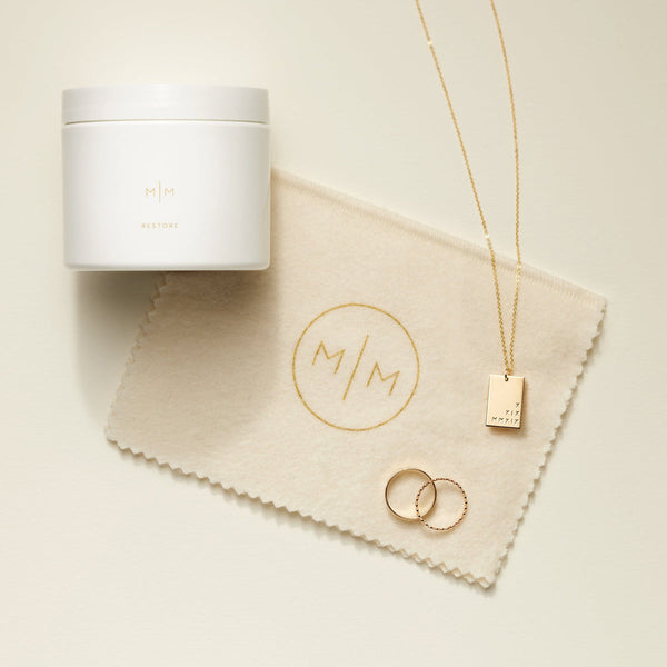 A white jar labeled 'RESTORE' on top of a beige polishing cloth, with a delicate gold rectangle pendant necklace and two gold rings placed to the right of it on a solid light beige background.