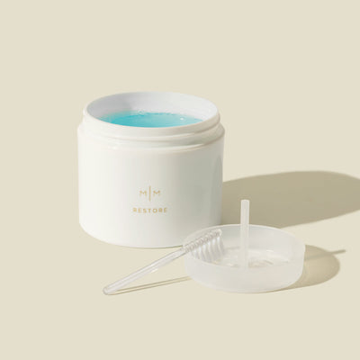 A white jar labeled 'RESTORE' with the top off showing a blue jewelry cleaning liquid, with a small clear cleaning brush and clear pan for setting jewelry pieces into the jar.