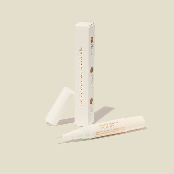 A tall, skinny white box standing vertically with an open white jewelry cleaner pen laying beside it. The box has the word 'RESTORE JEWELRY CLEANER PEN' down the front, and it is shown on a solid beige background.