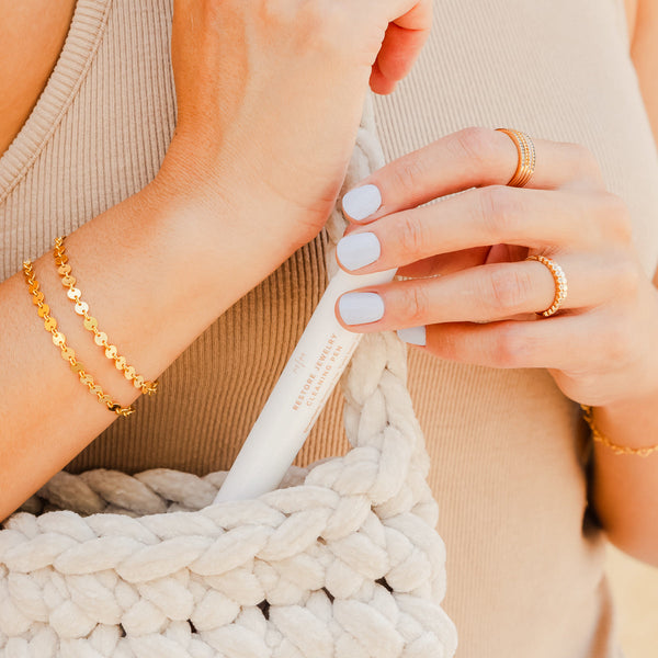 A person holding a white jewelry cleaner pen and placing it in a white braided texture purse. They are wearing a ribbed beige top and gold rings and bracelets.