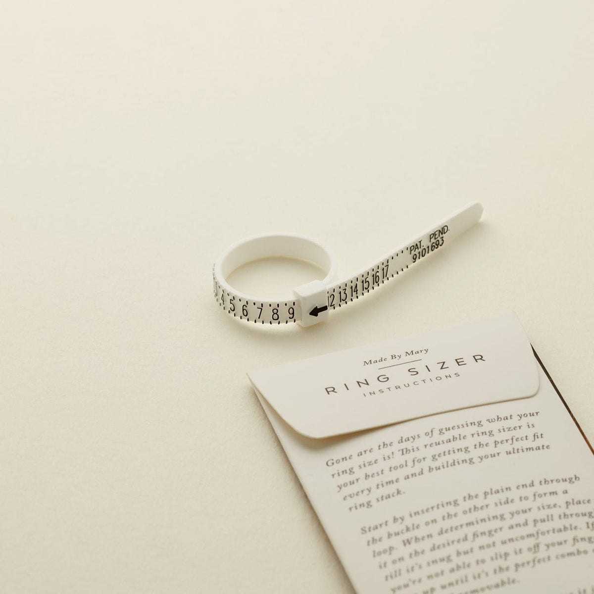 A white plastic zip-tie like ring sizer with black numbers, shown on a solid light beige surface.