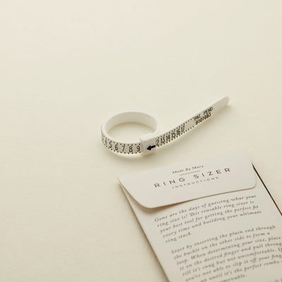 A white plastic zip-tie like ring sizer with black numbers, shown on a solid light beige surface.